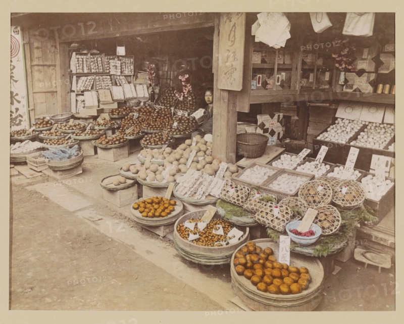 Display of foodstuffs at a shop, photograph, page, Japan, c. 1887 - in or before 1897, photographer, colorer (graphic design artist), paper, albumen print, hand coloring, height 187 mm x width 243 mm, Photo for page 123., shop, store, vegetables, A storefront with wooden beams and shelves is filled with round baskets and trays holding various fruits and goods, including piles of round fruits in different shades and textured items arranged on trays and in woven cages, with small rectangular markers placed among them; a seated person is partially visible behind a wooden post near the center, with a neutral facial expression; shelves and boxes line the back wall, some stacked and some displayed in open compartments, and a few vases with flowers sit on the shelves; the ground in front of the shop is a worn paved surface with scattered wooden boards and crates.