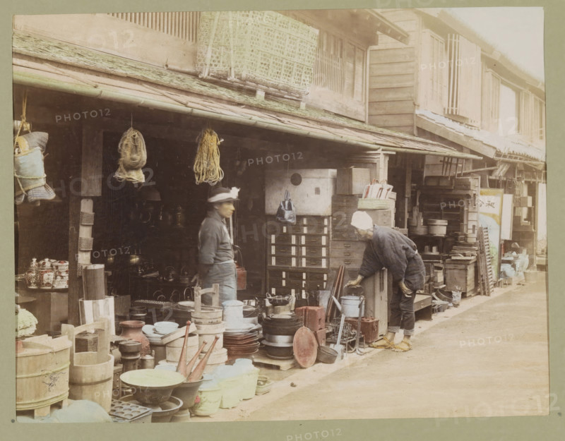 View of a shopping street, photograph, page, Japan, c. 1887 - in or before 1897, photographer, colorer (graphic design artist), paper, albumen print, hand coloring, photographie peinte, height 187 mm x width 243 mm, Photo for page 299., shop, store, street, shop-window, show-window, A street scene showing the front of a wooden shop with an overhanging tiled roof and various items displayed outside, including stacks of round and cylindrical containers, shallow bowls, trays, wooden buckets, metal pots, and other household wares arranged on the ground and on low platforms; two people stand near the shopfront, one facing sideways wearing a dark jacket and a hat with a brim, the other bending forward wearing a light head covering and dark clothing with sandals on their feet; hanging from the eaves are bundled ropes and wrapped objects, and a small bag is suspended near a column; the shop exterior features wooden shutters, small drawers and crates, and shelving filled with boxes and assorted...