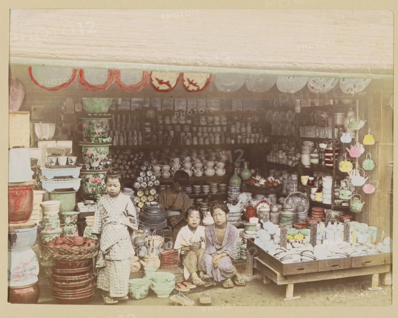 Shop with pottery, photograph, page, Japan, c. 1887 - in or before 1897, photographer, colorer (graphic design artist), paper, albumen print, hand coloring, photographie peinte, height 186 mm x width 243 mm, Photo for page 325., shop, store, container of ceramics: jar, jug, pot, vase, A storefront filled with stacked ceramics, bowls, jars, vases, plates, and teapots arranged on shelves, tables, and the floor, with patterned large pots and smaller cups in groups; hanging round decorative plates along the top edge and colorful hanging teapots on a rack to the right; a woman seated inside among the wares and three children in front of the display wearing patterned garments and sandals, one child standing while the others sit; baskets, trays, and stacks of dishes displayed on wooden platforms and crates, with various glazed finishes and floral designs visible.