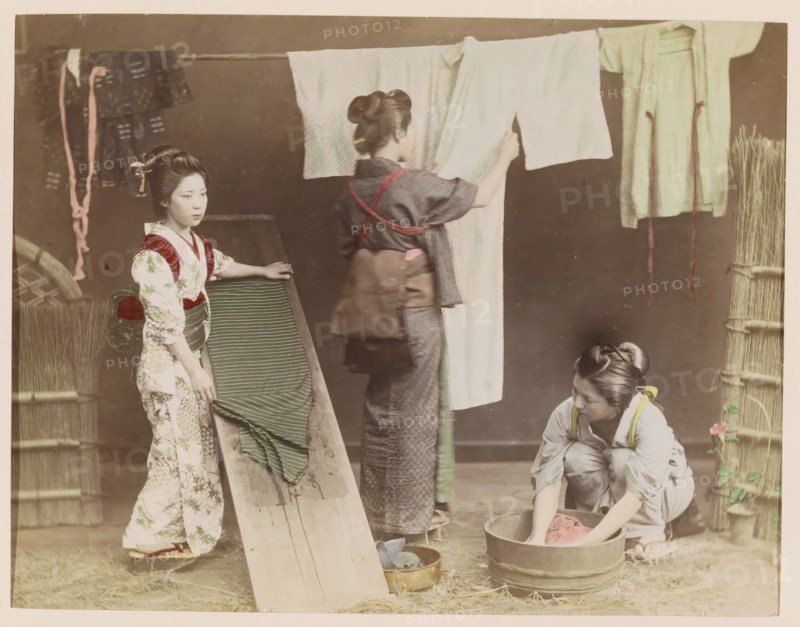 Three women washing kimonos, photograph, page, photographer: attributed to Kusakabe Kimbei, Japan, c. 1888 - in or before 1898, colorer (graphic design artist), Attributed to, paper, albumen print, hand coloring, photographie peinte, height 187 mm x width 244 mm, Photo for page 369., adult woman, dress, gown, wash house, home laundry, laundry room, Three women are engaged in household tasks in a muted indoor scene. One woman stands on the left wearing a patterned garment with a sash, her hair styled and adorned with a small hair ornament, and she rests a hand on a wooden incline draped with a striped cloth. A second woman stands center facing away, wearing a darker outfit with a tied sash and her hair in a bun, holding up a light-colored cloth that hangs from a horizontal pole. A third woman kneels on the right beside a wooden tub, sleeves rolled, working with a piece of cloth in the water, her hair tied back and a small floral detail near her. Hanging garments and ropes are visible on...