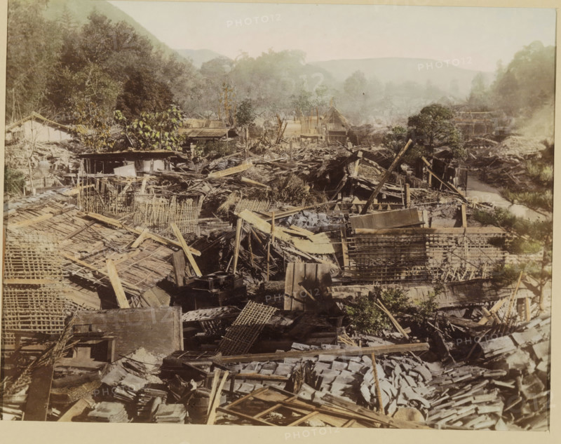 View of a village destroyed by an earthquake, photograph, page, Japan, c. 1888 - in or before 1898, photographer, colorer (graphic design artist), paper, albumen print, hand coloring, photographie peinte, height 186 mm x width 243 mm, Photo for page 375., landscape with ruins, consequences of an earthquake, A wide scene of an area filled with broken wooden structures and scattered planks, beams, and lattice panels, with piles of debris and roofing materials spread across the foreground and middle ground, interspersed with small trees and vegetation. In the background there are more collapsed wooden buildings and fragments of rooftops partially obscured by mist, with dense foliage and hills rising behind them. The overall palette shows muted earth tones and faded colors across the wreckage, vegetation, and distant hills.
