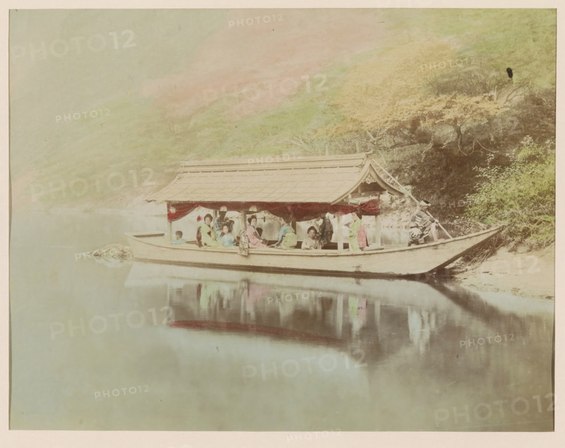 Houseboat, photograph, page, Japan, c. 1888 - in or before 1898, photographer, colorer (graphic design artist), paper, albumen print, hand coloring, photographie peinte, height 187 mm x width 244 mm, Photo for page 381., A long wooden boat with a roofed structure floats on calm water near a riverbank, with reflections visible on the water surface; several people sit inside the boat wearing traditional clothing in soft pastel colors, and red drapery hangs along the open sides of the roofed section; a person at the stern holds a pole and another person stands near the bow; behind the boat the bank shows trees and foliage with varied tones, and a gentle hillside rises in the background under a pale sky.