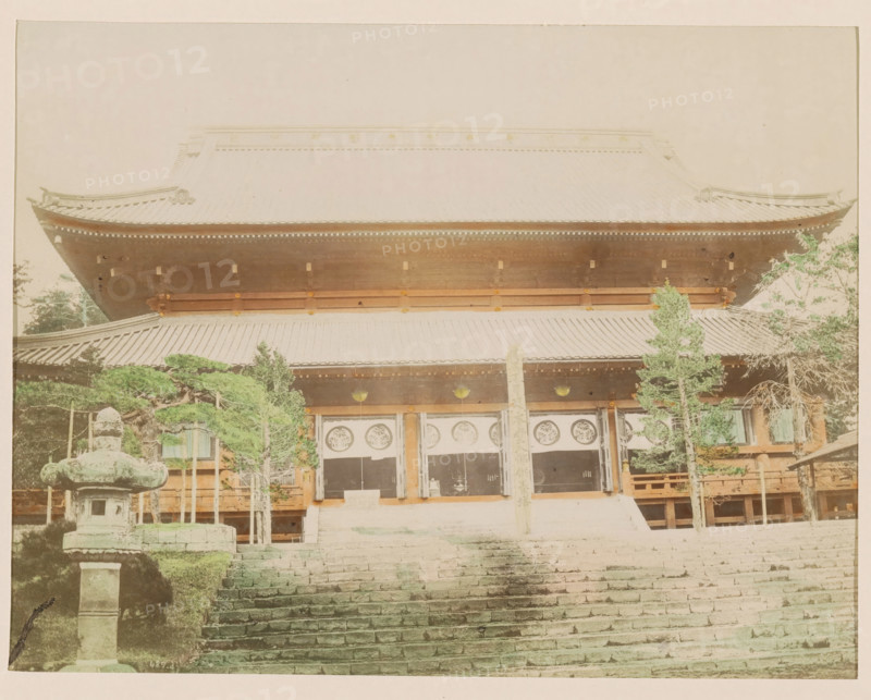 Front facade of the Manwangji Temple at Nikko, photograph, page, Nikko, Japan, c. 1888 - in or before 1898, photographer, colorer (graphic design artist), Kanto, paper, albumen print, hand coloring, photographie peinte, height 187 mm x width 242 mm, Photo for page 357., temple, shrine, Hinduism, Buddhism, Jainism, A large traditional wooden building with a tiered tiled roof fills the upper part of the image, showing ornate roof edges and eaves; beneath the roof a covered porch area with hanging circular decorations and wooden beams is visible, framed by wooden pillars and a central vertical post. Stone steps lead up toward the entrance in the foreground, flanked on the left by a weathered stone lantern and trimmed greenery, and on the right by several tall slender trees with green foliage. The scene has muted, slightly faded colors and a soft, even light across the structure and grounds.