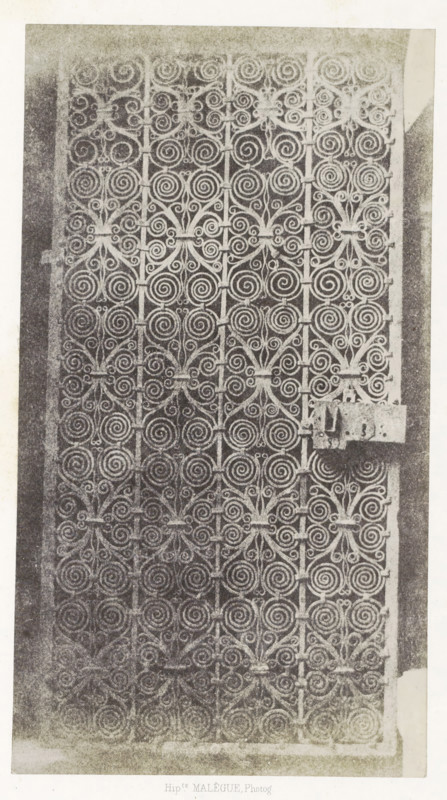 Porte en fer, Iron gate from the monastery of Le Puy-en-Velay, photograph, page, inscription: 'provenant du cloître de la Cathédrale du Puy.', number: 'PL. 13.', photographer: Hippolyte Malègue, France, c. 1850 - in or before 1857, paper, salted paper print, height 244 mm x width 135 mm, gate, entrance, ornament, circle and derived from circle, eg: guilloche, A vertically oriented image of an ornate metal gate filling most of the frame, showing a repeating pattern of spiral scrolls and floral-like motifs arranged in a grid, with vertical bars intersecting decorative elements and a rectangular metal latch attached on the right side; the gate is set within a lighter border and the overall image has a grainy, high-contrast texture.