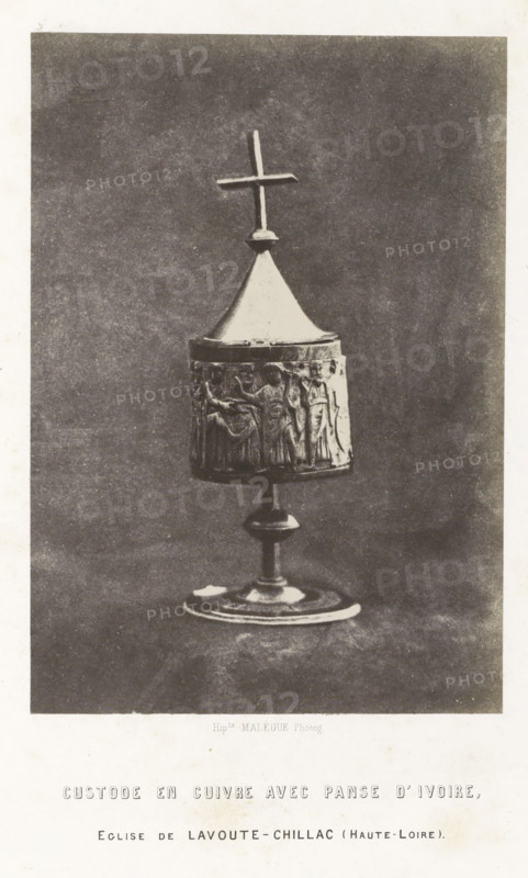 Ivory reliquary from Lavoûte-Chilhac, Custode en cuivre avec panse d'ivoire, photograph, page, inscription: 'eglise de Lavoute-Chillac (Haute-Loire).', number: 'PL. 16.', photographer: Hippolyte Malègue, France, c. 1850 - in or before 1857, Hippolyte Malègue, photographer, paper, salted paper print, height 195 mm x width 136 mm, relics (and reliquaries), A photographic image of a chalice-like object standing on a circular base with a central stem supporting a cylindrical body topped by a conical lid and a simple cross finial; the cylindrical section is decorated with a raised relief showing standing human figures in robes; the object appears metallic with a light-colored lid and darker relief band, set against a plain textured background and framed by a white border.