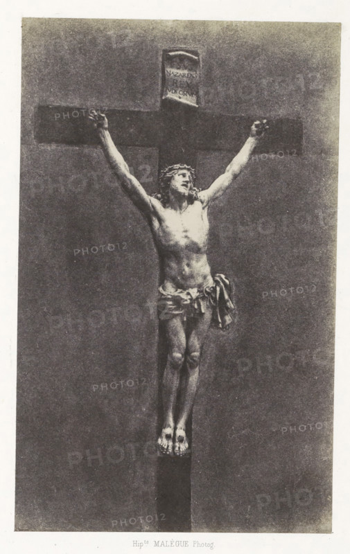Ivory crucifix from the cathedral of Le Puy, Christ en ivoire, photograph, page, inscription: 'appartenant a la cathédrale du Puy.', number: 'PL. 19.', photographer: Hippolyte Malègue, France, c. 1850 - in or before 1857, paper, salted paper print, height 200 mm x width 124 mm, sculpture, the crucifixion of Christ: Christ's death on the cross, Golgotha (Matthew 27:45-58, Mark 15:33-45, Luke 23:44-52, John 19:25-38), A monochrome image of a crucifix with a carved figure of a man affixed to a dark wooden cross, the figure shown full length with arms outstretched and hands positioned at the ends of the horizontal beam, a loincloth draped around the waist with fabric folds hanging at one side, a crown-like headpiece on the head and a tilted facial expression, a visible torso with muscular definition, legs together and feet resting on a small platform at the base of the vertical beam, a small rectangular plaque above the head attached to the top of the cross, and an even textured background...