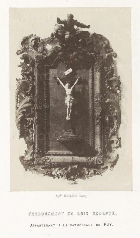Crucifix in wooden frame from the cathedral of Le Puy-en-Velay, Encadrement en bois sculpté, photograph, page, inscription: 'appartenant a la Cathédrale du Puy.', number: 'PL. 24.', photographer: Hippolyte Malègue, France, c. 1850 - in or before 1857, paper, salted paper print, height 188 mm x width 125 mm, God the Father represented as human being, usually with crown or tiara or scepter and, or globe, the crucifixion of Christ: Christ's death on the cross, Golgotha (Matthew 27:45-58, Mark 15:33-45, Luke 23:44-52, John 19:25-38), sculpture (relief, sculpture), A framed ornate relief occupies the center of the image, showing a sculpted figure on a cross suspended within a deep rectangular panel; the frame is richly carved with swirling acanthus-like foliage, shells, and clustered decorative elements, and a cherubic or bearded figure sits atop the frame with arms extended; the relief interior is darker than the surrounding carved border, and a shadow falls from the lower part of the cros...