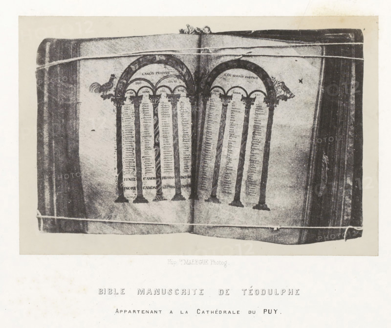 Bible manuscrite de Téodulphe, Bible from the cathedral of Le Puy-en-Velay, photograph, page, inscription: 'appartenant a la Cathédrale du Puy.', number: 'PL. 25.', photographer: Hippolyte Malègue, France, c. 1850 - in or before 1857, paper, salted paper print, height 136 mm x width 204 mm, Bible, An open book with visible pages showing decorative arches supported by columns, ornate capitals, and narrow vertical columns of small marks; small animal-like figures appear near the tops of the arches; the book edges and binding are visible along the sides, and light horizontal lines cross the image near the top and bottom margins.