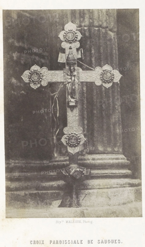Croix paroissiale de Saugues, Parochial cross from Saugues, photograph, page, number: 'PL. 3.', photographer: Hippolyte Malègue, France, c. 1850 - in or before 1857, paper, salted paper print, height 196 mm x width 129 mm, crucifix, personal devotion, Madonna: ie Mary with the Christ-child, A vertical photograph of an ornate metal cross mounted on a decorative stand placed in front of a textured columned background, the cross showing intricate floral and geometric patterns with rounded decorative ends, a small sculpted figure affixed at the center, and ribbons hanging from near the top, all rendered in a monochrome tone.