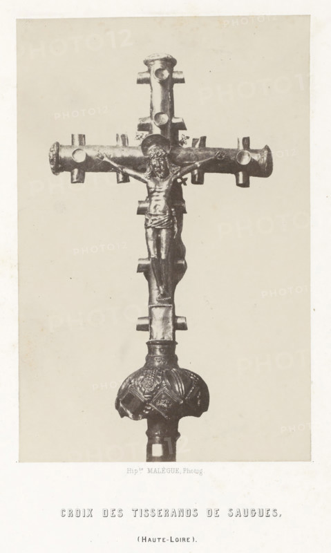 Cross of the weavers, from Saugues, Croix des tisserands de Saugues, photograph, page, number: 'PL. 5.', inscription: '(Haute-Loire).', photographer: Hippolyte Malègue, France, c. 1850 - in or before 1857, paper, salted paper print, height 202 mm x width 133 mm, crucifix, personal devotion, A vertical photograph of a metal crucifix mounted on a decorative stem, shown against a plain light background. The crucifix features a horizontal crossbar with rounded ends and small protruding cylindrical nodes, and a vertical shaft with matching nodes. A sculpted figure with a halo is affixed to the cross, depicted with outstretched arms, a draped lower garment, and visible facial and torso details. Below the cross, the stem widens into an ornamented rounded knob with embossed patterns and a textured surface, and the lower shaft continues downward out of frame. The image has a narrow light border around it.