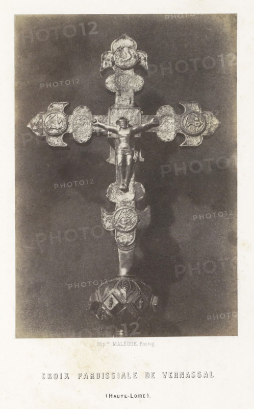 Parochial cross from Vernassal, Croix paroissiale de Vernassal, photograph, page, number: 'PL. 6.', inscription: '(Haute-Loire).', photographer: Hippolyte Malègue, France, c. 1850 - in or before 1857, paper, salted paper print, height 193 mm x width 133 mm, crucifix, personal devotion, A photographic image of an ornate metal cross with a central figure of a crucified person mounted on a dark rectangular background; the cross has decorative round medallions at the ends and along the shaft, each containing intricate embossed designs, and the base of the cross shows a rounded, patterned support.