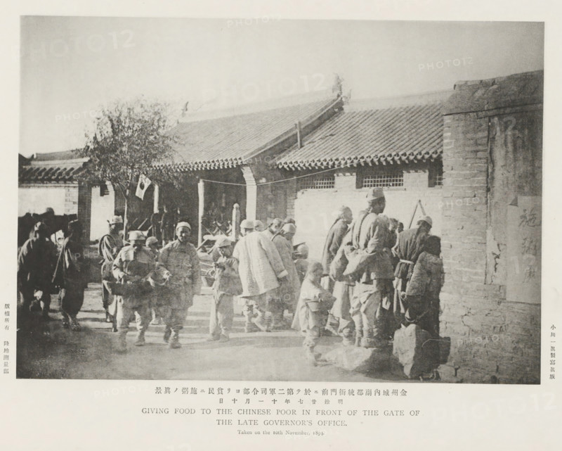 Giving food to the Chinese poor in front of the gate of the late governor's office, page, photomechanical print, inscription: 'The fall of Kinchow.', photographer: Ordnance Survey, Manchuria, cliché maker: Kazumasa Ogawa, Japan, 1894-11-10, Ordnance Survey, Kazumasa Ogawa, photographer, cliché maker, paper, collotype, photoprint, height 195 mm x width 275 mm, the soldier, the soldier's life, giving alms or other charity, eg: handing out food, facade (of house or building), A sepia-toned photograph showing a group of people gathered in front of a single-story building with a tiled sloping roof and decorative eaves; some individuals stand while others sit or bend, many wearing coats and hats, and a few carry bags or bundles; a tree with sparse foliage is visible near the building entrance, and a pillar and low wall appear at the right side of the image; shadows fall across the ground and the scene has a worn, textured appearance.