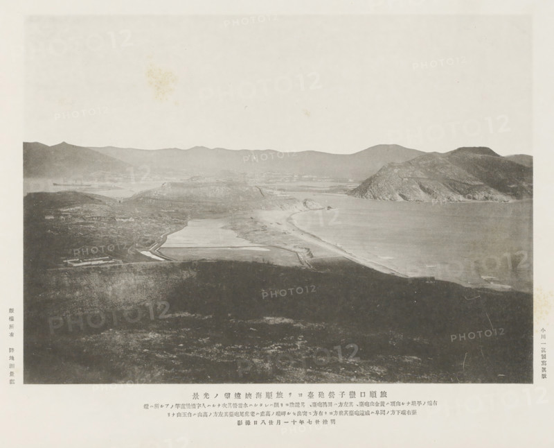 View of the Port Arthur straits from the Lantseying forts at Port Arthur, page, photomechanical print, inscription: 'The fall of Port Arthur.', inscription: 'The flat hill on the right end is, The high hill on the left is Peiyuh-shan.', photographer: Ordnance Survey, Manchuria, cliché maker: Kazumasa Ogawa, Japan, 1894-11-28, paper, collotype, photoprint, height 197 mm x width 272 mm, city-view in general, 'veduta', gulf, bay, A grayscale panoramic scene shows a coastal landscape with a wide beach curving along the shoreline next to calm water, rising rocky headlands and cliffs on the right, lower rolling hills and flat plains extending inland, and distant mountains forming a jagged horizon; the foreground consists of a darker, textured expanse of land with faint paths and irregular patches, while various lighter, rectangular shapes on the plain suggest constructed surfaces near the coast, and the sky above is pale and mostly empty with a few subtle blemishes.