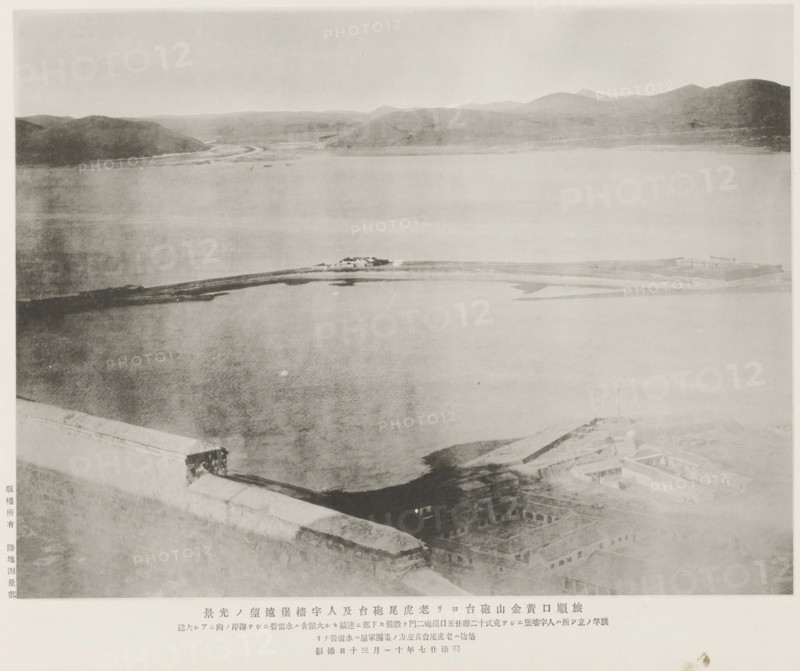 A distant view of the Old tiger's tail fort and the Jentse earworks from the Hwangkin hill forts, Port Arthur, page, photomechanical print, inscription: 'The place where the flag-staff stands is Jentse, to the left of which is a torpedo-station.', inscription: 'The fall of Port Arthur.', photographer: Ordnance Survey, Manchuria, cliché maker: Kazumasa Ogawa, Japan, 1894-11-30, Ordnance Survey, Kazumasa Ogawa, photographer, cliché maker, paper, collotype, photoprint, height 212 mm x width 278 mm, fortress, straits, narrows, A wide, pale grayscale view of a body of water bordered by distant hills under a light sky, with a narrow strip of land or causeway extending across the middle distance and small low structures clustered on it, the foreground showing a high stone or concrete wall edge and rooftop areas with rectangular openings and building outlines, and the overall image appearing faded and soft in contrast.