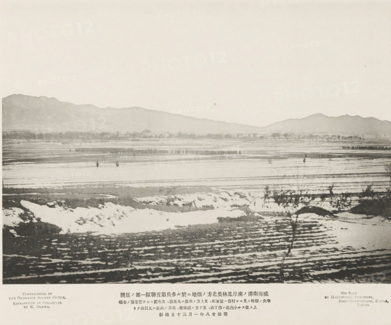 Extension of a portion of the fifth infantry regiment, in a field N. of Funglintsei, on the S. coast of Wei-Hai-Wei, page, photomechanical print, inscription: 'Expedition to Wei-hai-wei.', inscription: 'The village seen in the wood, The mountain on the right is Takowshan.', name: 'On Sale By Hakubundo, Publisher, Nishi-Ogawamachi, Kanda, Tokyo.', photographer: Ordnance Survey, Shandong, cliché maker: Kazumasa Ogawa, Japan, 1895-01-30, paper, collotype, photoprint, height 203 mm x width 281 mm, army 'en route', A wide horizontal photograph showing a flat icy or snowy plain in the foreground with irregular patches of darker exposed ground and shallow ruts or rows, a few thin bare shrubs or small bushes casting faint shadows, a middle ground of more level frozen surface with scattered small dark upright figures and a distant linear group of tiny dark shapes, and a background of low rolling mountains stretching across the image beneath a pale, mostly empty sky.