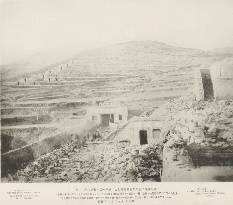 The rear defenses of the hill to the N. of the Tseszetai forts, on the W. Coast of Wei-Hai-Wei, page, photomechanical print, inscription: 'The road running by the telegraph post, to the left of the forts.', inscription: 'Expedition to Wei-hai-wei.', photographer: Ordnance Survey, Shandong, cliché maker: Kazumasa Ogawa, Japan, 1895-02-06, paper, collotype, photoprint, height 222 mm x width 286 mm, low hill country, fortress, defense, siege, A wide, pale landscape showing terraced hills with layered, horizontal ledges stretching across the scene, scattered with small rectangular openings in the slopes and rough rocky surfaces; in the foreground there are ruins and rubble leading to two stone buildings with arched doorways and flat roofs, one building partly buried and another framed by a taller stone wall and narrow steps, with a few indistinct human figures near the structures and a tall slender pole rising from the lower terraces.