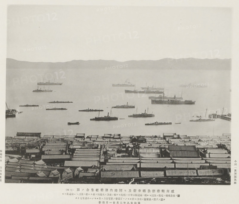 A view of Wei-Hai-Wei harbour, Liukung Island and its town, and the vessels in harbour, page, photomechanical print, inscription: 'Expedition to Wei-Hai-Wei.', inscription: 'Sunken on the right side, to the left are also official residences.', photographer: Ordnance Survey, Shandong, cliché maker: Kazumasa Ogawa, Japan, 1895-02-21, paper, collotype, photoprint, height 207 mm x width 263 mm, harbour, ships (in general), A wide waterfront scene shows a foreground of densely packed roofed structures with tiled and corrugated surfaces arranged in rows, a middle area of calm water dotted with various steamships and smaller boats at different distances, and a distant low mountain range along the horizon under a pale sky.