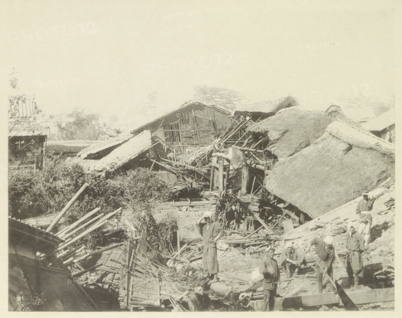 Destroyed houses in Biwajima after an earthquake, page, photomechanical print, Japan, cliché maker: Kazumasa Ogawa, after 1891-10-28 - in or before 1892, photographer, paper, collotype, photoprint, height 222 mm x width 285 mm, ruin of a dwelling, house, castle, consequences of an earthquake, Mino–Owari Earthquake 1891, A sepia-toned scene showing collapsed and damaged thatched and tiled roofs with exposed wooden beams and broken structural elements, scattered debris and wooden poles across the ground, a group of people wearing simple clothing and head coverings standing and sitting among the rubble, one person standing near the center with a hand raised to their head, vegetation and trees visible behind the damaged buildings, and partial remnants of remaining roof sections and scaffolding throughout the image.