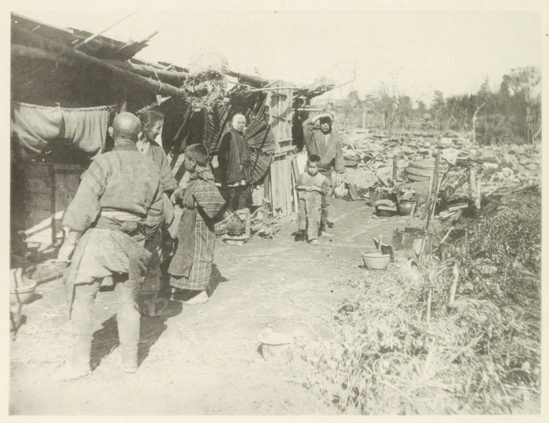 Temporary housing in Japan after an earthquake, page, photomechanical print, Japan, cliché maker: Kazumasa Ogawa, after 1891-10-28 - in or before 1892, photographer, paper, collotype, photoprint, height 198 mm x width 261 mm, housing, consequences of an earthquake, Mino–Owari earthquake 1891, A faded black-and-white scene showing several people standing and walking along a dirt path beside a row of makeshift wooden structures with thatched roofing and hanging cloths, with one person facing away from the camera in simple clothing and others grouped nearby in traditional garments, a child standing near the front looking forward, another person shielding their eyes with a hand, various household items and containers scattered along the path and near the structures, a large round woven object leaning against a wall, debris and more containers extending into the background toward a treeline under a pale sky.