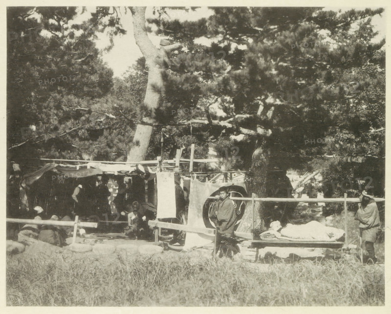 University field hospital in Japan after an earthquake, page, photomechanical print, Japan, cliché maker: Kazumasa Ogawa, after 1891-10-28 - in or before 1892, photographer, paper, collotype, photoprint, height 224 mm x width 283 mm, medical aid and nursing of the sick and wounded in a field-hospital, consequences of an earthquake, Mino–Owari earthquake 1891, A shaded outdoor scene shows large trees with dense branches and foliage filling the upper area and casting shadows below where a simple wooden structure with hanging cloth panels and draped fabric stands. Several people are visible around and within the structure, some seated and some standing, wearing layered garments and cloaks; one person stands near the front next to a circular patterned cloth, and another person sits or reclines on a low bench covered with folded textiles. A low wooden fence runs across the middle of the image, and grassy ground occupies the foreground. The overall image is monochrome with high contrast betw...