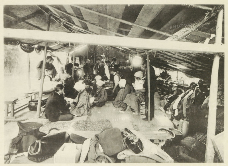 University field hospital in Japan after an earthquake, page, photomechanical print, photographer: Nakamura, Japan, cliché maker: Kazumasa Ogawa, after 1891-10-28 - in or before 1892, paper, collotype, photoprint, height 191 mm x width 264 mm, medical aid and nursing of the sick and wounded in a field-hospital, consequences of an earthquake, Mino–Owari earthquake 1891, A crowded interior scene under a sloped roof supported by poles, with a group of people seated and standing along a central aisle, many wearing head coverings and layered clothing, some seated on low stools or the floor and others standing and leaning; fabric, bags, and folded items are piled in the foreground, while benches, containers, and hanging items line the sides; the roof shows exposed beams and woven material, and light filters through openings creating bright patches and contrasts across the space.