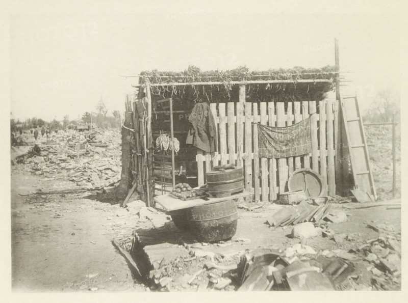 Temporary store in Japan after an earthquake, page, photomechanical print, Japan, cliché maker: Kazumasa Ogawa, after 1891-10-28 - in or before 1892, photographer, paper, collotype, photoprint, height 195 mm x width 275 mm, shop, store, ruin of a building, architecture, consequences of an earthquake, Mino–Owari Earthquake 1891, A weathered wooden shack with a thatched roof made of bundled vegetation stands at the edge of a debris-strewn dirt path, its front composed of vertical wooden slats and an open doorway revealing interior shelving and hanging rope; a coat hangs from a post and a patterned cloth is draped across part of the slatted wall, while various containers including stacked barrels, a round basin, baskets, and a board with a pile of round produce rest in front; a tall leaning ladder and scattered planks and rubble surround the structure, and a distant line of trees and indistinct figures can be seen along the horizon.