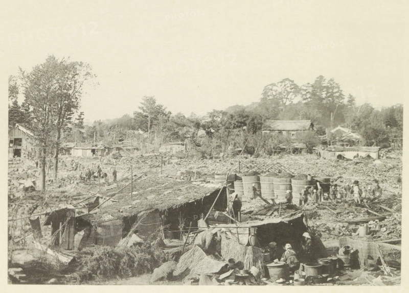 Destroyed buildings in Kasamatsu after an earthquake, page, photomechanical print, Kasamatsu, cliché maker: Kazumasa Ogawa, Japan, after 1891-10-28 - in or before 1892, photographer, Chubu, Gifu, paper, collotype, photoprint, height 211 mm x width 285 mm, ruin of a building, architecture, consequences of an earthquake, Mino–Owari earthquake 1891, A wide, desaturated scene shows an outdoor area cluttered with makeshift structures, thatch roofs, and scattered debris extending into a background of trees and low buildings; large cylindrical wooden tubs are grouped near the center right with small human figures near them, and more people are visible throughout the scene engaged in various activities; trees rise on the left and in the distance, and several simple houses and sheds are interspersed among vegetation; in the foreground there are bundles of straw, containers, baskets, and tools arranged near the huts, with poles and ropes stretching across parts of the image.