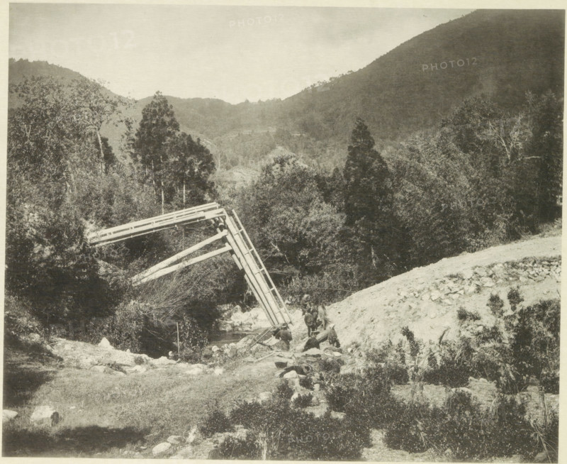 Destroyed bridge in the Neo Valley in Japan after an earthquake, page, photomechanical print, Japan, cliché maker: Kazumasa Ogawa, after 1891-10-28 - in or before 1892, photographer, paper, collotype, photoprint, height 227 mm x width 279 mm, bridge, dale, valley, consequences of an earthquake, Mino–Owari Earthquake 1891, A sepia-toned landscape showing a valley flanked by tree-covered hills and a distant ridge, with dense foliage of tall trees and shrubs in the midground and foreground. A wooden trestle structure leans diagonally over a small stream, with angled beams and rail-like planks visible. On a rocky slope near the stream a group of people are gathered, some standing and some seated, with packs and equipment on the ground nearby. The foreground shows grassy and rocky terrain with scattered stones and low vegetation. The sky above is pale and mostly clear.
