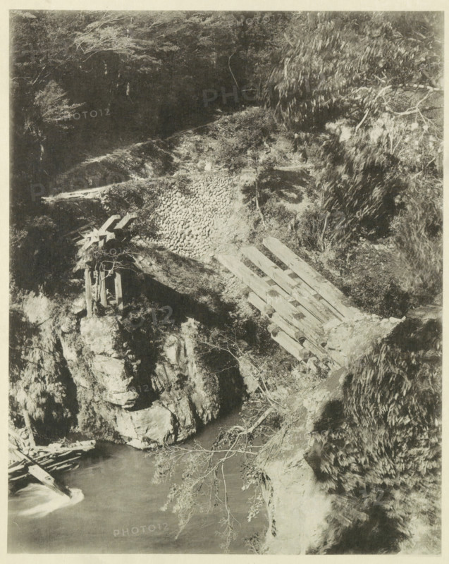 View of a destroyed bridge in the Neo Valley in Japan after an earthquake, page, photomechanical print, Japan, cliché maker: Kazumasa Ogawa, after 1891-10-28 - in or before 1892, photographer, paper, collotype, photoprint, height 283 mm x width 225 mm, bridge, dale, valley, consequences of an earthquake, Mino–Owari earthquake 1891, A monochrome photograph of a rocky riverside scene showing a wooden footbridge or planks laid across a steep bank, with stacked stones forming a retaining area beside the planks, rough rock outcrops, and a vertical wooden support structure perched on a ledge; water flows at the bottom with visible surface movement, fallen branches lie near the water, and vegetation including leafy trees and shrubs frames the scene with overhanging branches and textured foliage.