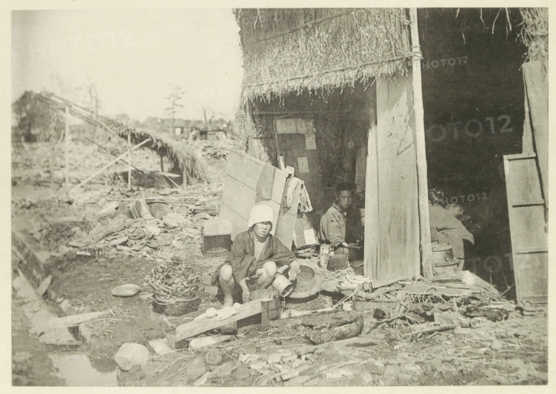 Temporary house in a village in Japan destroyed by an earthquake, page, photomechanical print, Japan, cliché maker: Kazumasa Ogawa, after 1891-10-28 - in or before 1892, photographer, paper, collotype, photoprint, height 196 mm x width 276 mm, ruin of a dwelling, house, castle, consequences of an earthquake, Mino–Owari Earthquake 1891, A faded photograph shows a thatched structure with a partially open doorway and a dark interior, while debris and scattered objects lie in front of it; a person wearing a light head covering and patterned clothing is crouched near a low wooden plank, holding a container, and another person is seated inside the doorway partially visible among household items and containers, with various baskets, bowls, planks, and bundles arranged on the ground around them.