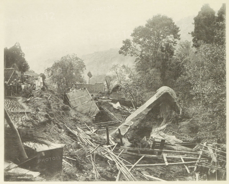 Destroyed houses in the Neo Valley in Japan after an earthquake, page, photomechanical print, Japan, cliché maker: Kazumasa Ogawa, after 1891-10-28 - in or before 1892, photographer, paper, collotype, photoprint, height 230 mm x width 288 mm, ruin of a building, architecture, consequences of an earthquake, Mino–Owari Earthquake 1891, A sepia-toned scene showing a collapsed rural area with broken wooden beams, planks, and ladders scattered across uneven ground, overturned thatched roofs and fragmented structures among mud and debris, dense trees and bushes rising on both sides with a large tree prominent on the right, a narrow muddy path or trench winding through the wreckage, a small group of people standing near upright remnants and stacked materials at the left edge, and distant hazy hills visible through the mist above the treeline.