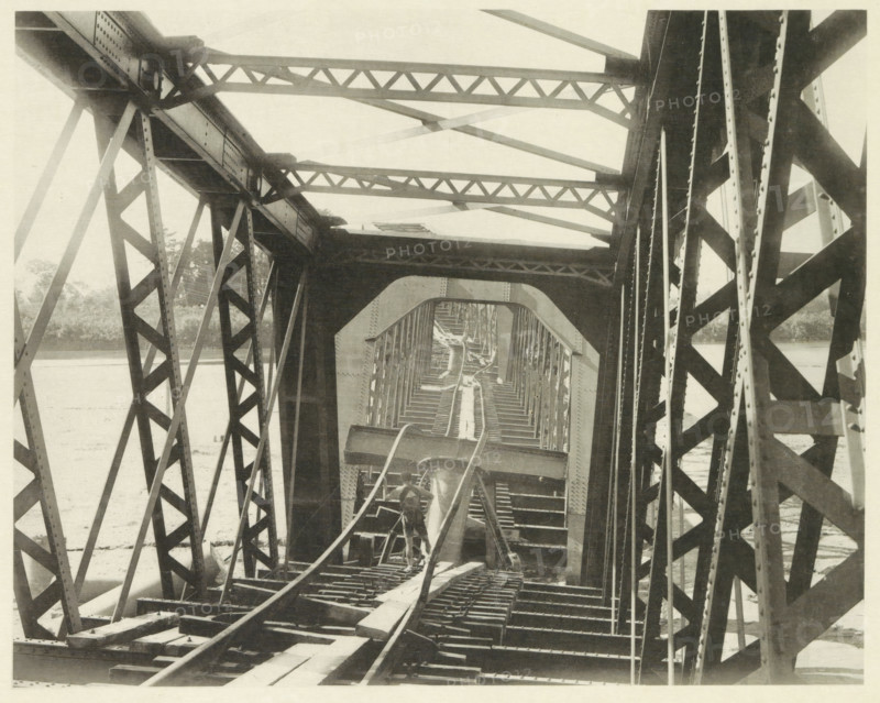 Collapsed railway bridge over the Nagara River in Japan after an earthquake, page, photomechanical print, Japan, cliché maker: Kazumasa Ogawa, after 1891-10-28 - in or before 1892, photographer, paper, collotype, photoprint, height 210 mm x width 266 mm, railway, tramway, rack railway, bridge, consequences of an earthquake, Mino–Owari earthquake 1891, A view through a metal truss bridge showing intersecting diagonal and horizontal beams forming repeating triangular patterns, riveted plates and girders, a central walkway with wooden planks and parallel rails, a man standing on the structure near the foreground facing away, layered arch-like openings framing the bridge interior, water visible beyond the bridge on either side, and distant vegetation along the far bank.