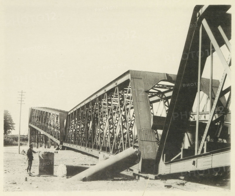 Collapsed railway bridge over the Nagara River in Japan after an earthquake, page, photomechanical print, Japan, cliché maker: Kazumasa Ogawa, after 1891-10-28 - in or before 1892, photographer, paper, collotype, photoprint, height 230 mm x width 278 mm, railway, tramway, rack railway, bridge, consequences of an earthquake, Mino–Owari earthquake 1891, A large steel truss bridge spans from the foreground into the distance with visible riveted girders and latticework, angled support beams and curved arch elements, and a section of heavy diagonal braces in the right foreground; a sandy ground area beneath the bridge shows scattered debris and a cylindrical concrete block with a person standing beside it holding a bicycle or similar two-wheeled object; a utility pole rises at the left edge and a small tree is visible near it, while the sky above is empty.