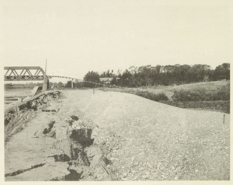 View of a collapsed railway bridge over the Nagara River in Japan after an earthquake, page, photomechanical print, Japan, cliché maker: Kazumasa Ogawa, after 1891-10-28 - in or before 1892, photographer, paper, collotype, photoprint, height 218 mm x width 281 mm, railway, tramway, rack railway, bridge, consequences of an earthquake, Mino–Owari earthquake 1891, A wide gravelly expanse fills the foreground, showing a surface of loose stones and a narrow stick lying across the rocks, with an eroded dirt path and jagged exposed soil along the left edge. In the middle distance a slender pole rises from the ground near a low concrete support, and a trussed metal bridge section extends from the left toward the center, with a gently curved beam running toward a wooded area. Beyond the bridge, a line of trees and dense shrubbery stretches across the scene, with grassy embankments sloping down toward the gravel. The sky above is pale and empty.