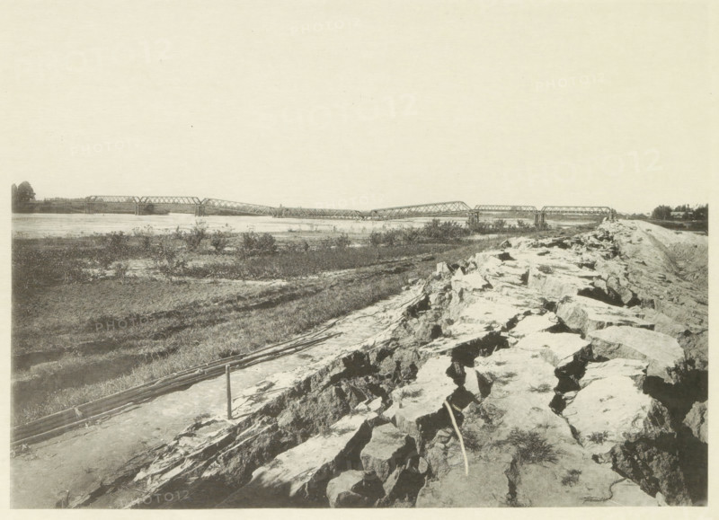View of a collapsed railway bridge over the Nagara River in Japan after an earthquake, page, photomechanical print, Japan, cliché maker: Kazumasa Ogawa, after 1891-10-28 - in or before 1892, photographer, paper, collotype, photoprint, height 204 mm x width 282 mm, railway, tramway, rack railway, bridge, consequences of an earthquake, Mino–Owari earthquake 1891, A wide riverside scene with a long metal truss bridge spanning across the water in the distance, supported by piers and showing multiple connected spans; the near foreground shows a rocky embankment and uneven stone surface with scattered grasses and a narrow path or cleared strip alongside it; midground areas of low vegetation and patches of bare soil extend toward the riverbank; on the far shore there are trees and small clusters of foliage; the sky above is pale and mostly empty.