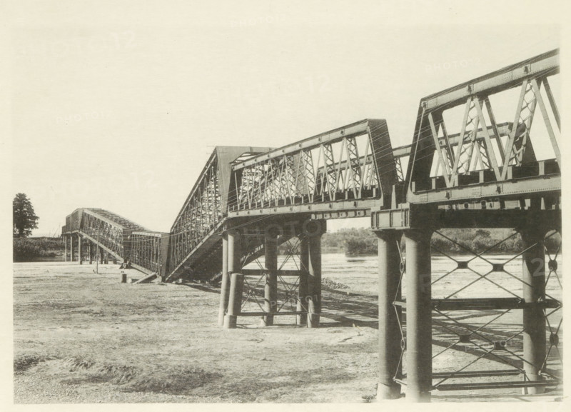 Collapsed railway bridge over the Nagara River in Japan after an earthquake, page, photomechanical print, Japan, cliché maker: Kazumasa Ogawa, after 1891-10-28 - in or before 1892, photographer, paper, collotype, photoprint, height 197 mm x width 287 mm, railway, tramway, rack railway, bridge, consequences of an earthquake, Mino–Owari earthquake 1891, A long metal truss bridge spans from the foreground to the background, showing repeating triangular framework and large vertical supports rising from the ground and water; the bridge sections slope and connect at different angles, with visible rivets and girders, and shadowed undersides above a mostly dry, textured riverbed; a solitary tree stands near the far bank, and distant foliage lines the horizon beneath a pale, empty sky.