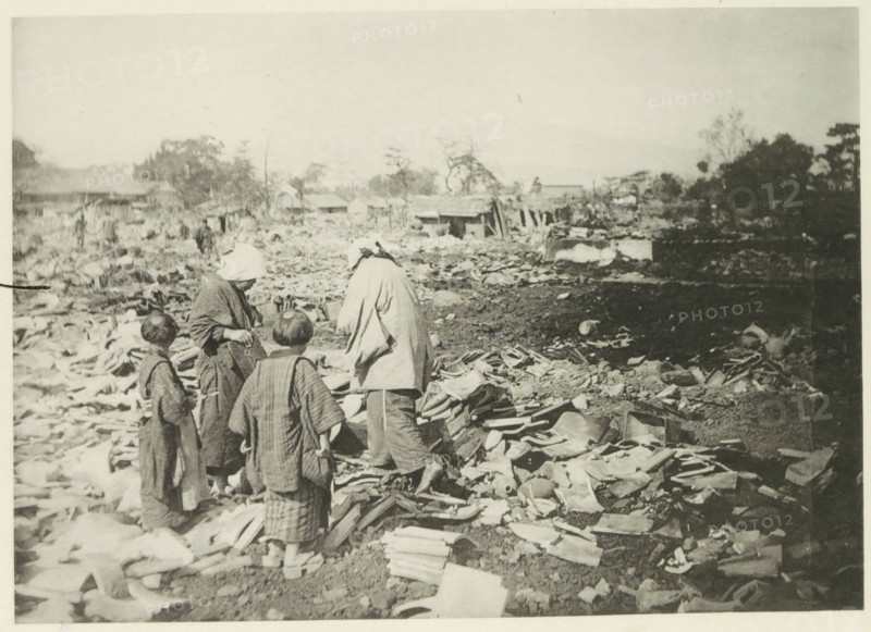 Two women with children stand among the destruction in Japan after an earthquake, page, photomechanical print, Japan, cliché maker: Kazumasa Ogawa, after 1891-10-28 - in or before 1892, photographer, paper, collotype, light print, height 181 mm x width 254 mm, ruin of a dwelling, house, castle, consequences of an earthquake, Mino–Owari Earthquake 1891, A sepia-toned photograph shows four people standing among a wide expanse of broken tiles and debris scattered across the ground, with three appearing to wear layered, loose garments and one wearing a head covering. The foreground is filled with irregular fragments of roofing tiles and curved ceramic pieces piled and strewn across dirt. In the midground, low wooden structures and scattered materials extend across the scene, with a few indistinct figures farther back. The background contains sparse trees, clusters of taller foliage, and more rubble extending toward a pale, empty sky.
