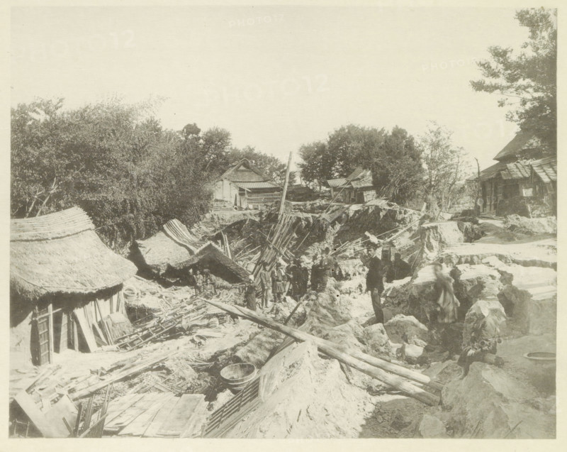 Houses destroyed by an earthquake in Japan, page, photomechanical print, Japan, cliché maker: Kazumasa Ogawa, after 1891-10-28 - in or before 1892, photographer, paper, collotype, photoprint, height 221 mm x width 276 mm, ruin of a dwelling, house, castle, consequences of an earthquake, Mino–Owari Earthquake 1891, A sepia photograph shows a rural scene with uneven ground and scattered wooden beams and planks. Thatched roofs and simple wooden structures appear along the left and right edges, and a small building with a sloped roof sits amid trees in the middle distance. Trees and foliage occupy much of the upper left and right areas. Several people stand or sit among the debris and earth, some near a shallow trench and others on rocky ground. Woven baskets and round containers lie on the ground, and a leaning pole rises from the center of the scene. The overall view includes rough pathways and piles of earth and construction materials.