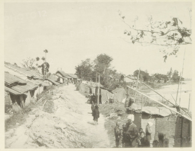Temporary housing along the Biwajima River in Japan, page, photomechanical print, Japan, cliché maker: Kazumasa Ogawa, after 1891-10-28 - in or before 1892, photographer, paper, collotype, photoprint, height 223 mm x width 287 mm, ruin of a dwelling, house, castle, housing, consequences of an earthquake, Mino–Owari Earthquake 1891, A dirt path runs through a village lined with simple thatched and tiled roofs on one side and fabric-covered stalls and woven fences on the other, with several people walking and standing along the path and clustered near the foreground; trees and tall slender palms rise behind the houses toward the center, while a branch with sparse leaves frames the upper right, and a river or body of water is visible beyond the fences on the right.