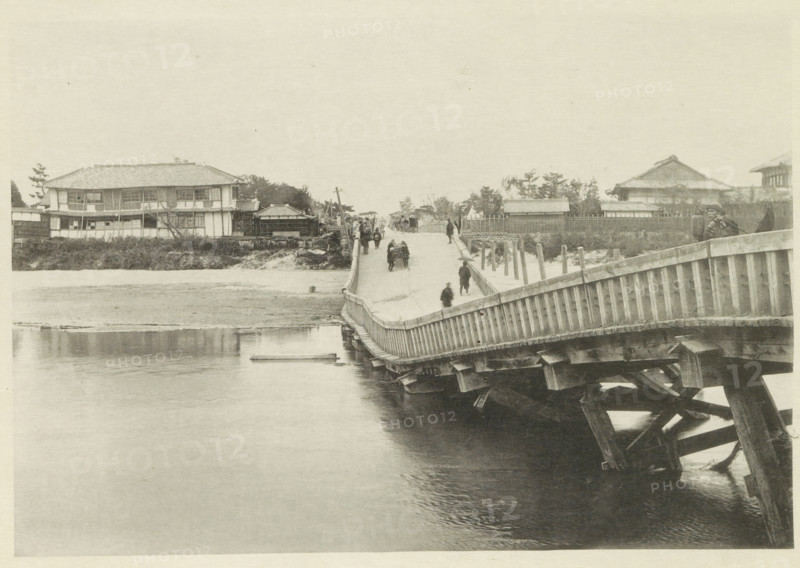 Collapsed bridge over the Biwajima River in Japan, page, photomechanical print, Japan, cliché maker: Kazumasa Ogawa, after 1891-10-28 - in or before 1892, photographer, paper, collotype, photoprint, height 196 mm x width 286 mm, bridge, consequences of an earthquake, Mino–Owari earthquake 1891, A wide wooden bridge extends from the foreground toward the distant shore, showing its slatted railing and supporting beams visible over water beneath, with several people walking along its length and a few standing near the far end; on the left bank there are low buildings with tiled roofs and rectangular windows set behind a grassy embankment, while the right bank features fenced yards, trees, and additional tiled-roof structures, and calm water fills the lower portion of the scene reflecting parts of the bridge.
