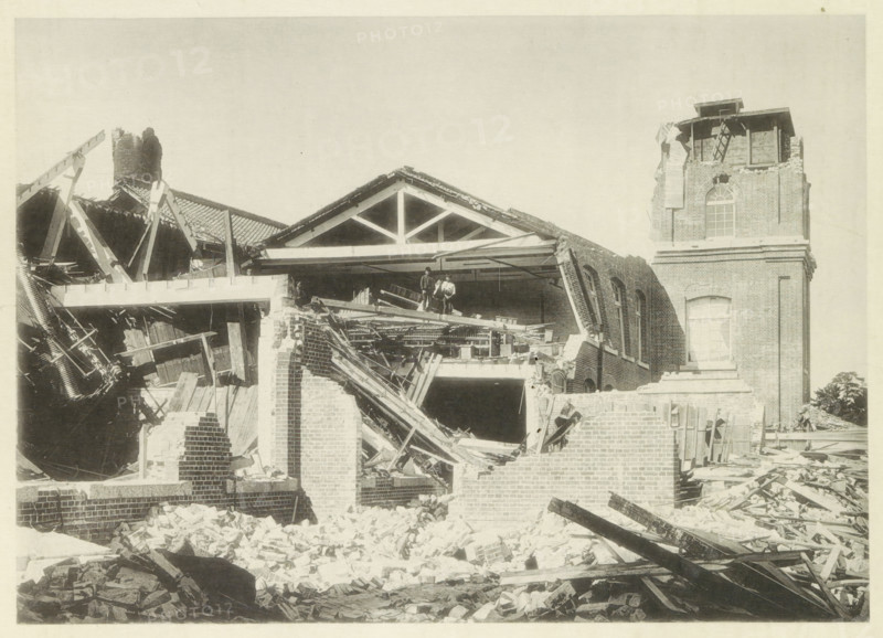 Collapsed cotton mill in Nagoya, page, photomechanical print, Nagoya, cliché maker: Kazumasa Ogawa, Japan, after 1891-10-28 - in or before 1892, photographer, Chubu, paper, collotype, photoprint, height 207 mm x width 290 mm, collapsing building, consequences of an earthquake, Mino–Owari earthquake 1891, A partially collapsed brick building with a gabled roof structure exposed, showing broken wooden beams, scattered bricks, and rubble in the foreground, two figures standing on a damaged interior floor amid debris, an intact tower section with arched windows and a damaged upper portion, leaning walls and exposed rafters, and piles of timber and masonry strewn across the site.