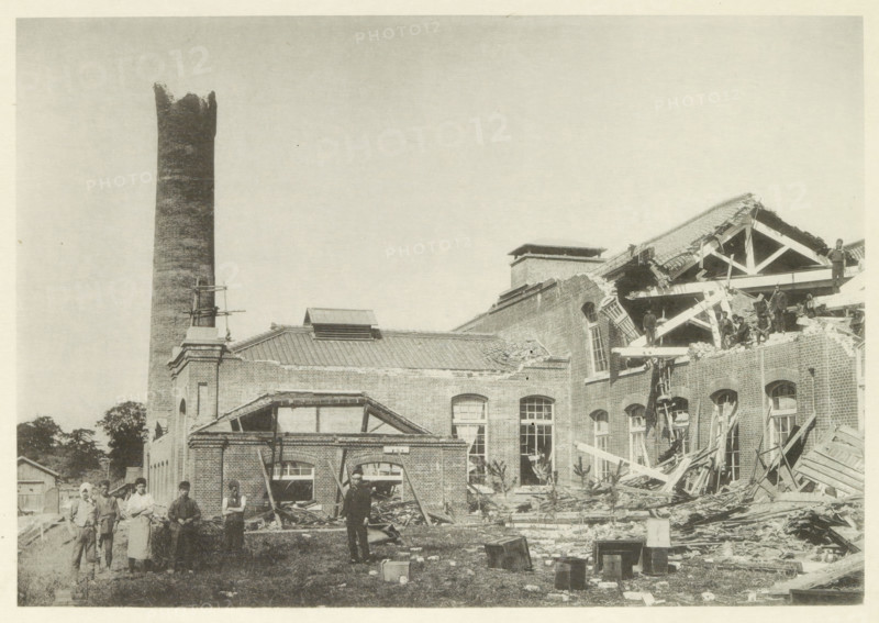 Collapsed cotton mill in Nagoya, page, photomechanical print, Nagoya, cliché maker: Kazumasa Ogawa, Japan, in or after 1891-10-28 - in or before 1892, photographer, Chubu, paper, collotype, photoprint, height 202 mm x width 289 mm, collapsing building, consequences of an earthquake, Mino–Owari earthquake 1891, A large brick building with a tall cylindrical chimney stands against a pale sky, part of the structure showing a collapsed roof and exposed wooden beams and rafters; broken brickwork, scattered timber, and debris cover the ground in front of the building, where several people stand among the rubble, some on the ruined upper floor and others at ground level; windows and arched openings in the brick facade are visible, along with a small pitched roof section and a rectangular rooftop vent; a few wooden crates and loose materials lie on the grassy area in the foreground.