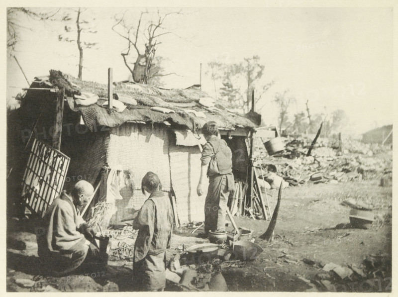Two women and a man in front of a temporary home in Japan, page, photomechanical print, Japan, cliché maker: Kazumasa Ogawa, after 1891-10-28 - in or before 1892, photographer, paper, collotype, photoprint, height 213 mm x width 290 mm, housing, consequences of an earthquake, Mino–Owari earthquake 1891, A sepia-toned outdoor scene shows a makeshift shelter with a woven fabric covering and rough materials on its roof, supported by vertical poles and surrounded by scattered tools and containers. Several people stand or sit near the shelter, wearing simple clothing and facing the shelter or each other. Pots, pans, buckets, and other domestic items are arranged on the ground in front of the shelter. Leafless trees rise behind the shelter and a landscape of rubble and uneven ground extends into the background.