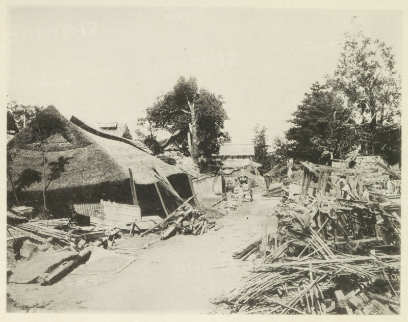 Destroyed buildings along the road to Gifu, page, photomechanical print, Japan, cliché maker: Kazumasa Ogawa, after 1891-10-28 - in or before 1892, photographer, paper, collotype, photoprint, height 218 mm x width 280 mm, public road, ruin of a building, architecture, consequences of an earthquake, Mino–Owari earthquake 1891, A wide dirt path runs through a scene of wooden debris and thatched roofs, with piles of planks and broken timber scattered along both sides; on the left a large thatched structure with irregular roof edges and nearby smaller wooden crates and frameworks sit amid rubble, while on the right stacks of wooden beams and lattice-like pieces form a cluttered foreground leading toward rudimentary wooden scaffolding and frames; in the middle distance a pair of cartlike objects with rounded fronts stand on the path flanked by more damaged wooden materials, and behind them a cluster of trees with leafy canopies rises above small rooflines and simple structures.