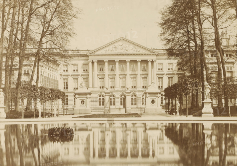 View of the facade of the Palace of the Nation in Brussels, Palais de la Nation, Bruxelles, photograph, page, name: '(Phot. de Brand frères, rue de la Madeleine, 32.)', photographer: Brand Frères, Brussels, c. 1862 - c. 1872, paper, albumen print, height 150 mm x width 205 mm, Photo on page 3., Lower House, Parliament, Chamber, pond, pool, façade (of house or building), Palace of the Nation, A sepia-toned photograph of a neoclassical building with a columned central facade and a triangular pediment decorated with sculpted reliefs, framed by tall leafless trees on both sides, a formal garden with trimmed hedges and several statues on ornate pedestals in the foreground, a reflective rectangular pool mirroring the building and trees, rows of evenly spaced windows and decorative balustrades along the roofline, and gated entrances at the base of the central section.