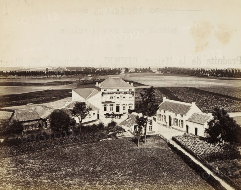 View of the Hôtel du Musée te Waterloo, Waterloo, photograph, page, name: '(Phot. de Brand frères, rue de la Madeleine, 32.)', photographer: Brand Frères, c. 1862 - c. 1872, paper, albumen print, height 150 mm x width 187 mm, Photo on page 15., rural housing, eg country-house, villa, cottage, façade (or house or building), A wide sepia-toned panoramic view shows a rural landscape with several white buildings clustered near the center, including a prominent multi-story house with a pitched roof and decorative facade, flanked by smaller houses with tiled roofs. In front of the main house a horse-drawn carriage is parked near a courtyard area and a few trees stand nearby. Surrounding the buildings are cultivated fields and garden plots with visible rows of plants, separated by narrow paths and low fences. A line of trees and hedgerows runs across the middle distance, and beyond them open flat farmland stretches to the horizon under a pale sky. The foreground consists of a large textured f...