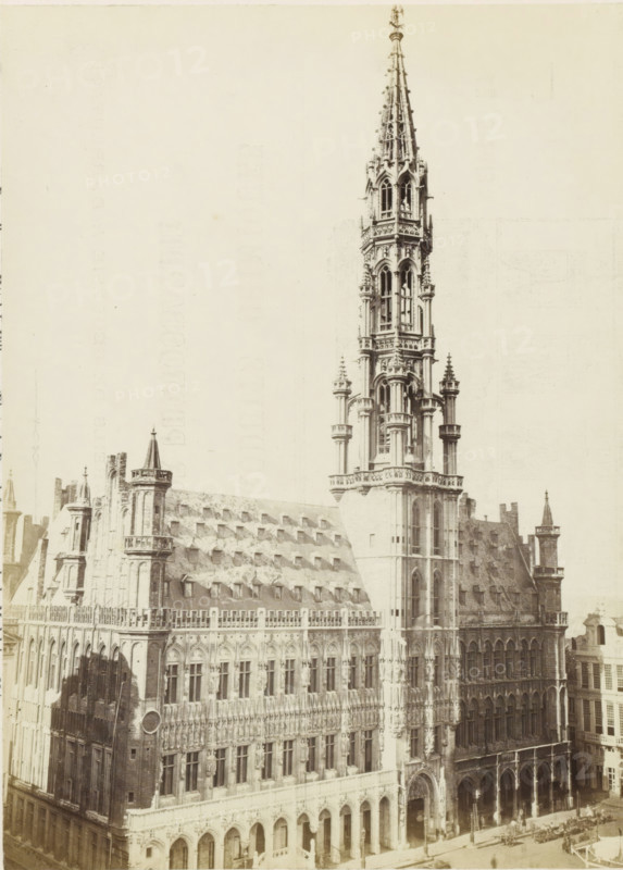 Town Hall of Brussels, Bruxelles - Hôtel-de-ville, photograph, page, name: '(Phot. de Brand frères, rue de la Madeleine, 32.)', photographer: Brand Frères, Brussels, c. 1862 - c. 1872, paper, albumen print, height 205 mm x width 151 mm, Photo on page 19., town hall, façade (of house or building), Stadhuis (Brussels), A large ornate stone building fills the image, featuring a tall, intricately detailed spire rising above a multi-story façade. The façade displays rows of arched windows and decorative stonework, including columns, statues, and carved patterns. One section shows a steep roof with a series of small dormer windows, and several turrets and pinnacles punctuate the roofline. At street level there are large archways and entrances, and a few small figures and horse-drawn vehicles are visible near the building's base. The photograph has a pale, aged tone with high contrast between the building details and an empty sky.