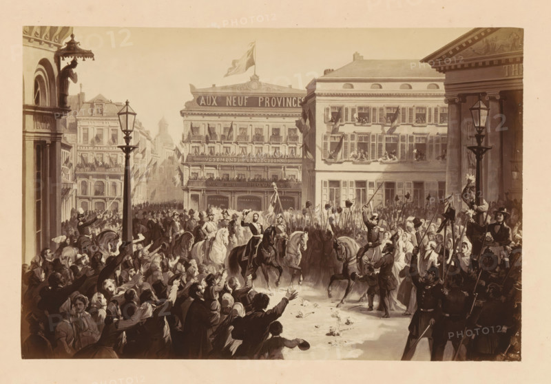 Photo reproduction of a design of the arrival of Léopold II of Belgium at Place de la Monnaie in Brussels during his inauguration on December 17, 1865, Entrée de SM Léopold 11. - le 17 Décembre 1865. Place de la Monnaie, photograph, reproduction, This photo is part of a cover with fourteen photos., photographer: Ghémar Frères, Brussels, after design by anonymous, c. 1861 - in or before 1866, paper, albumen print, albumendruk, height 167 mm x width 250 mm, Person, Leopold II (king of the Belgians), king, ceremonial celebration of installation of a ruler, Period, 1865-12-17, A crowded street scene with a dense throng of people lining both sides of a broad avenue, many reaching or waving toward a group of mounted figures at the center who ride dark and light horses and wear distinctive uniforms and hats; behind the riders a column of foot soldiers holds long poles with hats or banners aloft, and several individuals carry a tall flag; multi-story buildings with rows of windows and balconie...