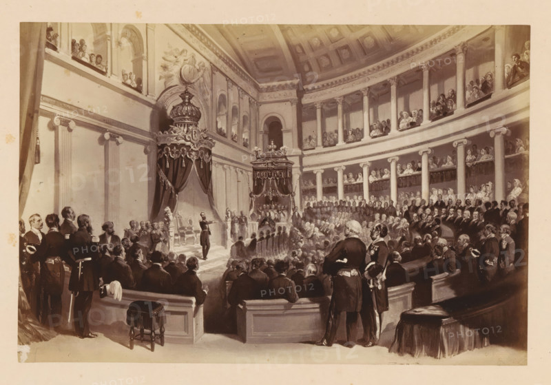 Photographic reproduction of a design of Léopold II of Belgium taking the oath during his inauguration on December 17, 1865 in Brussels, Presentation de Serment de SM Léopold 11, photograph, reproduction, This photo is part of a cover with fourteen photos., inscription: '17 Décembre 1865., Chambre des Représentants Bruxelles', photographer: Ghémar Frères, Brussels, after design by anonymous, c. 1861 - in or before 1866, paper, albumen print, height 166 mm x width 249 mm, Person, Leopold II (King of the Belgians), king, installation of a ruler, Period, 1865-12-17, A large, sepia-toned interior scene of an ornate, domed hall filled with many formally dressed people seated and standing on tiered balconies and in the main floor area, with tall columns and decorative molding lining the curved walls. At the focal point a raised platform is framed by heavy draped canopies topped with ornate crowns and decorative finials, flanked by sculpted pedestals and a throne-like chair. Numerous men in d...