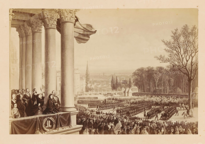 Hommage a la Garde Civique - Revue prize du balcon du Palais du Roi, Photo reproduction of a design of a tribute to the militia during the inauguration of Léopold II of Belgium in Brussels, photograph, reproduction, This photo is part of a cover with fourteen photos., inscription: 'Place des Palais.', photographer: Ghémar Frères, Brussels, after design by anonymous, c. 1861 - in or before 1866, paper, albumen print, albumendruk, height 167 mm x width 250 mm, Person, Leopold II (king of the Belgians), king, guard duty, keeping sentry, Period, 1865-12-17, A large balcony with ornate columns occupies the left foreground, a draped cloth hanging over its railing and a wreath attached to the fabric, and a group of people stand and sit on the balcony wearing formal clothing and cloaks, some facing outward toward the scene below. Beyond the balcony a procession of uniformed figures marches in organized formations across a broad open plaza filled with spectators, with mounted riders at the fron...